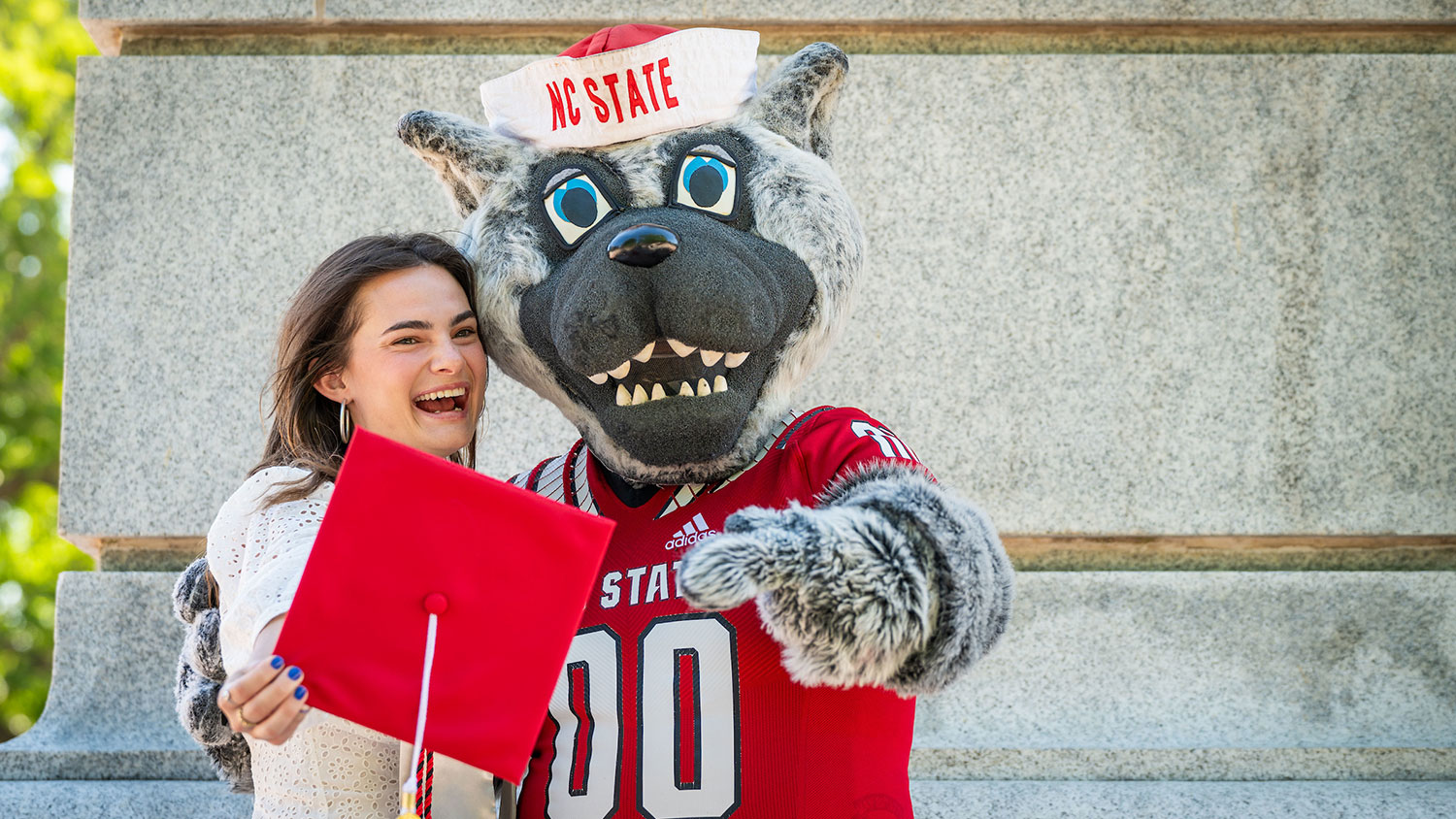 A student stands with Mr. Wuf while holding her graduation cap.