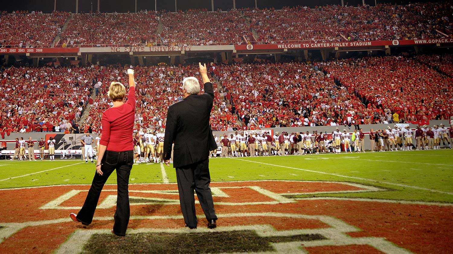 Chancellor Randy Woodson and his wife, Susan Woodson, stand on the football field surrounded by cheering Wolfpack fans.