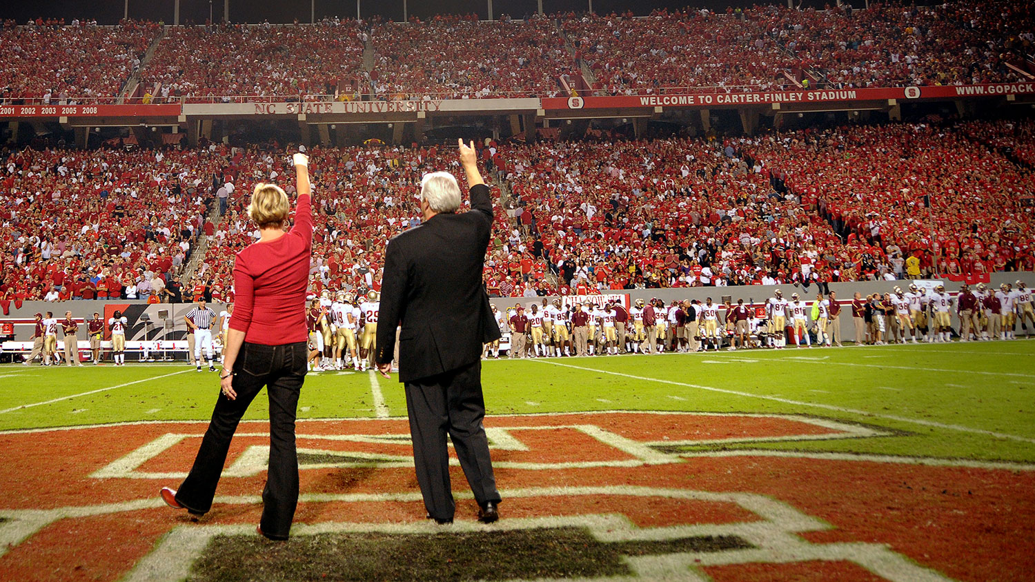 Chancellor Randy Woodson and his wife, Susan Woodson, stand on the football field surrounded by cheering Wolfpack fans.